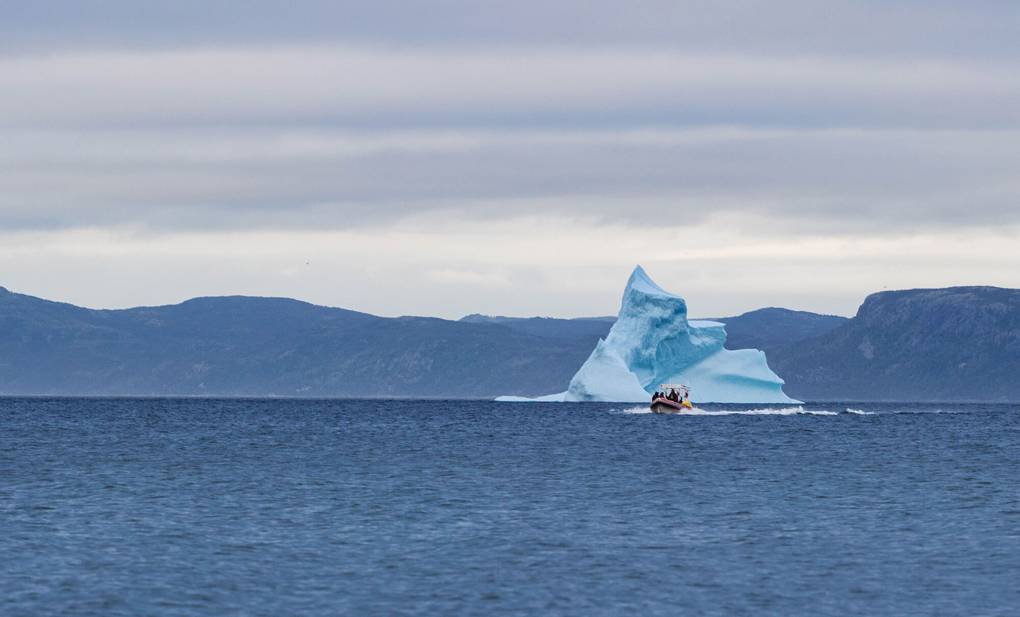 A Large Iceberg On The Horizon In Newfoundland With A Small Boat Driving Around, Conception Bay, Newfoundland And Labrador, Canada