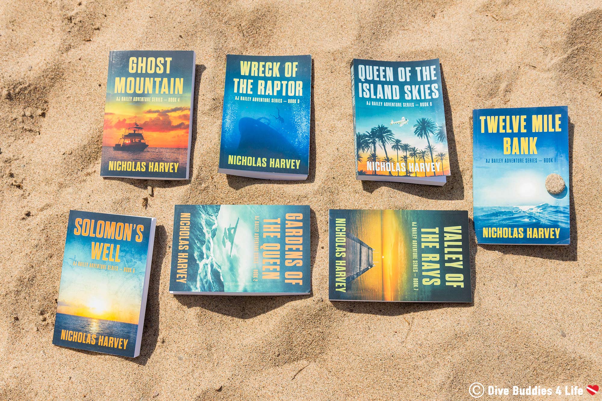 The Collection Of AJ Bailey Scuba Diving Novels Layed Out In The Sand Of A Beach